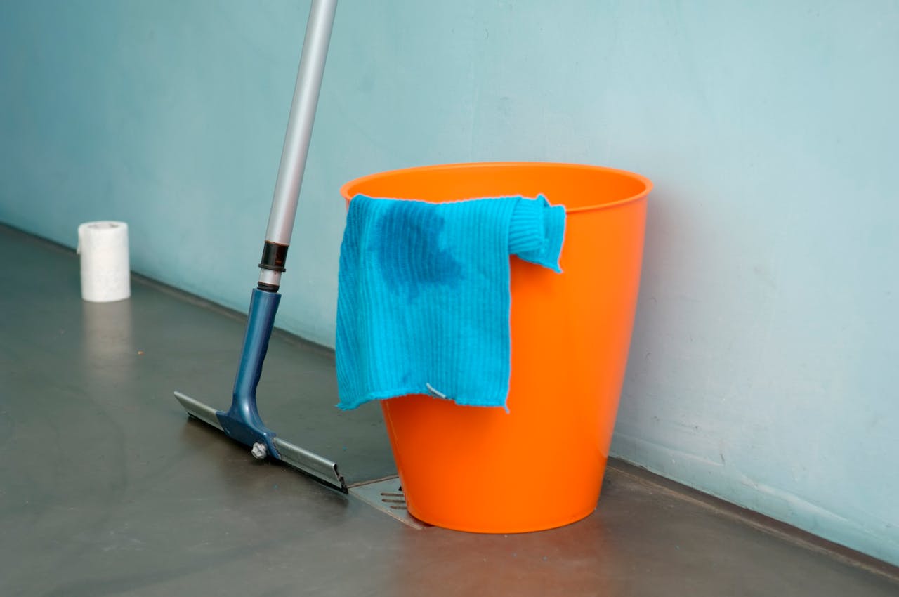 hero-img-02 Close-up of cleaning supplies including orange bucket, mop, and blue rag indoors.